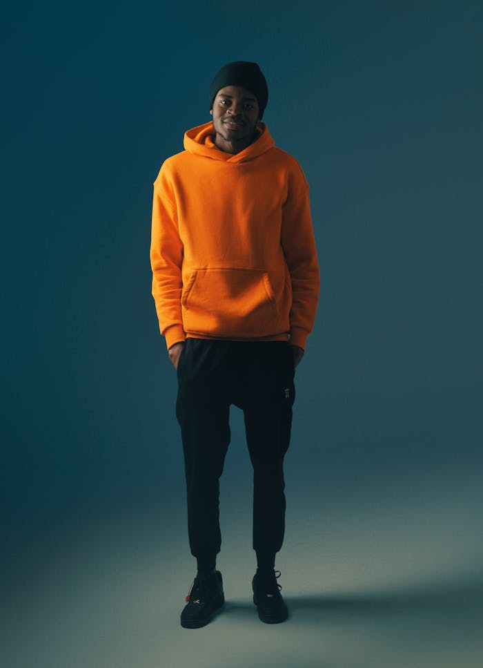 Young male model in orange hoodie posing against a blue backdrop in a studio setting.